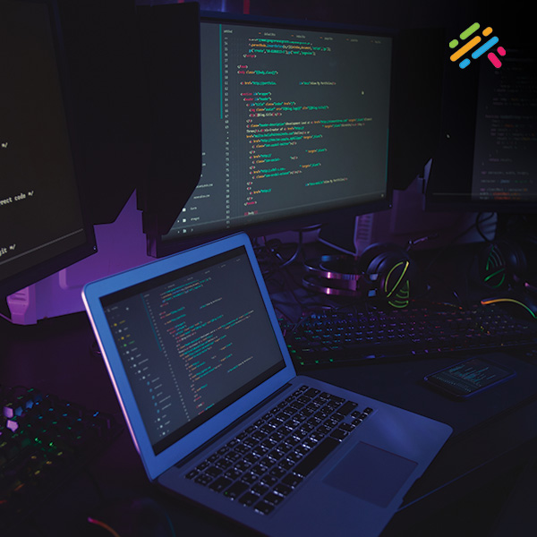 A person's hands typing on a laptop displaying lines of code, with two additional monitors glowing softly in a dim room.