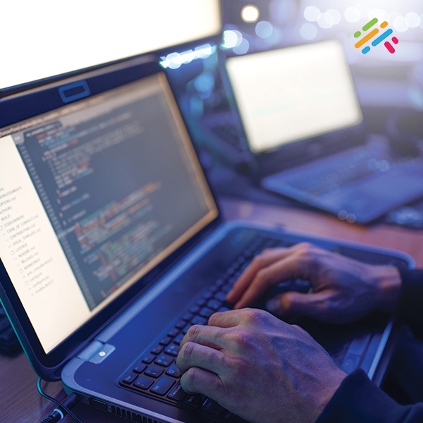 A close-up of hands typing on a laptop keyboard displaying programming code, with additional monitors glowing softly in the background.