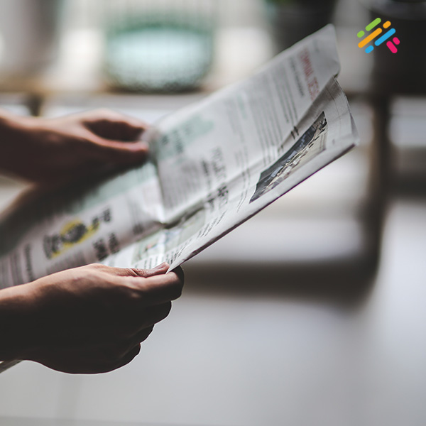 A close-up of two hands unfolding a printed newspaper at a home table, with a softly blurred living-room background.